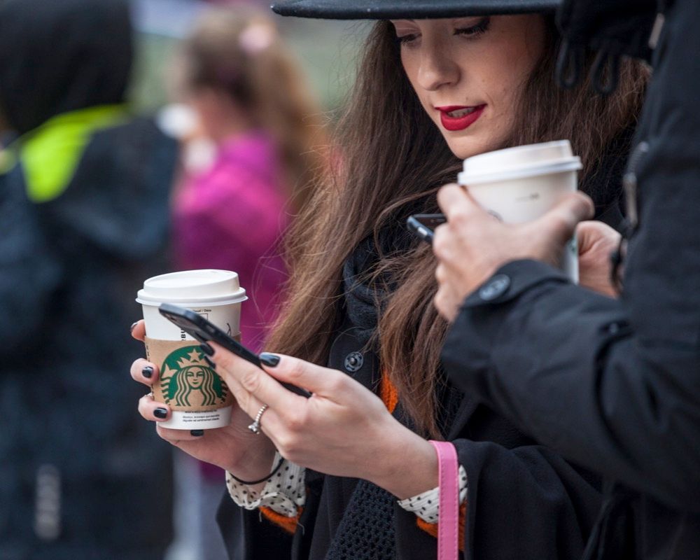 Picture of a young woman holding her smartphone while drinking a coffee in a cardboard coffee cup with the logo of Starbucks.