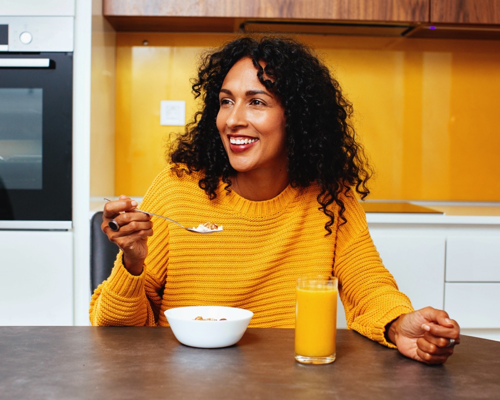 Portrait of a mid woman with black curly hair smiling while eating breakfast cereal in kitchen stock photo